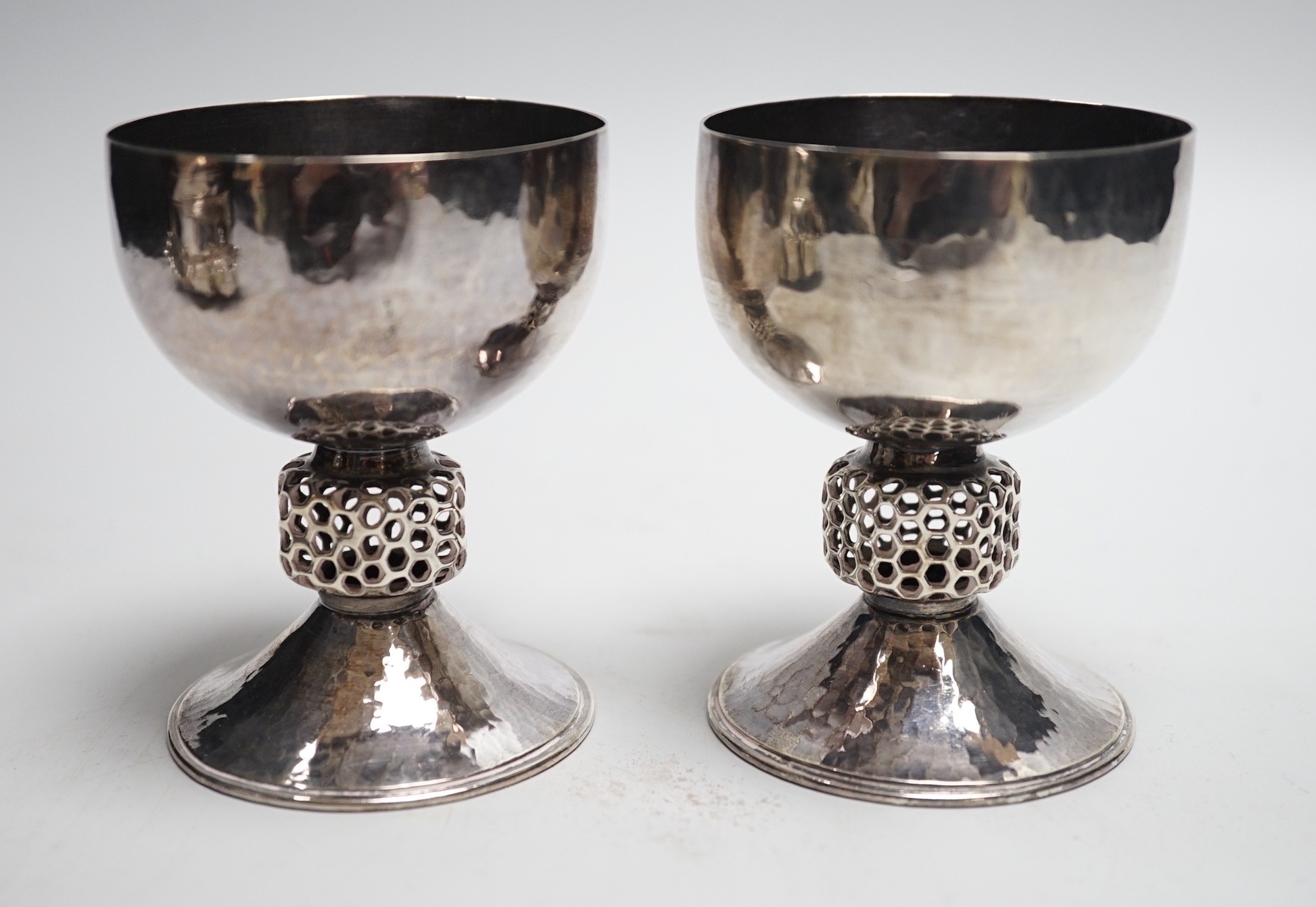 A stylish pair of Elizabeth II Arts & Crafts style planished silver pedestal cups, with reticulated stems, by Oliver Bailey, London, 1973, height 93mm, 7.5oz.